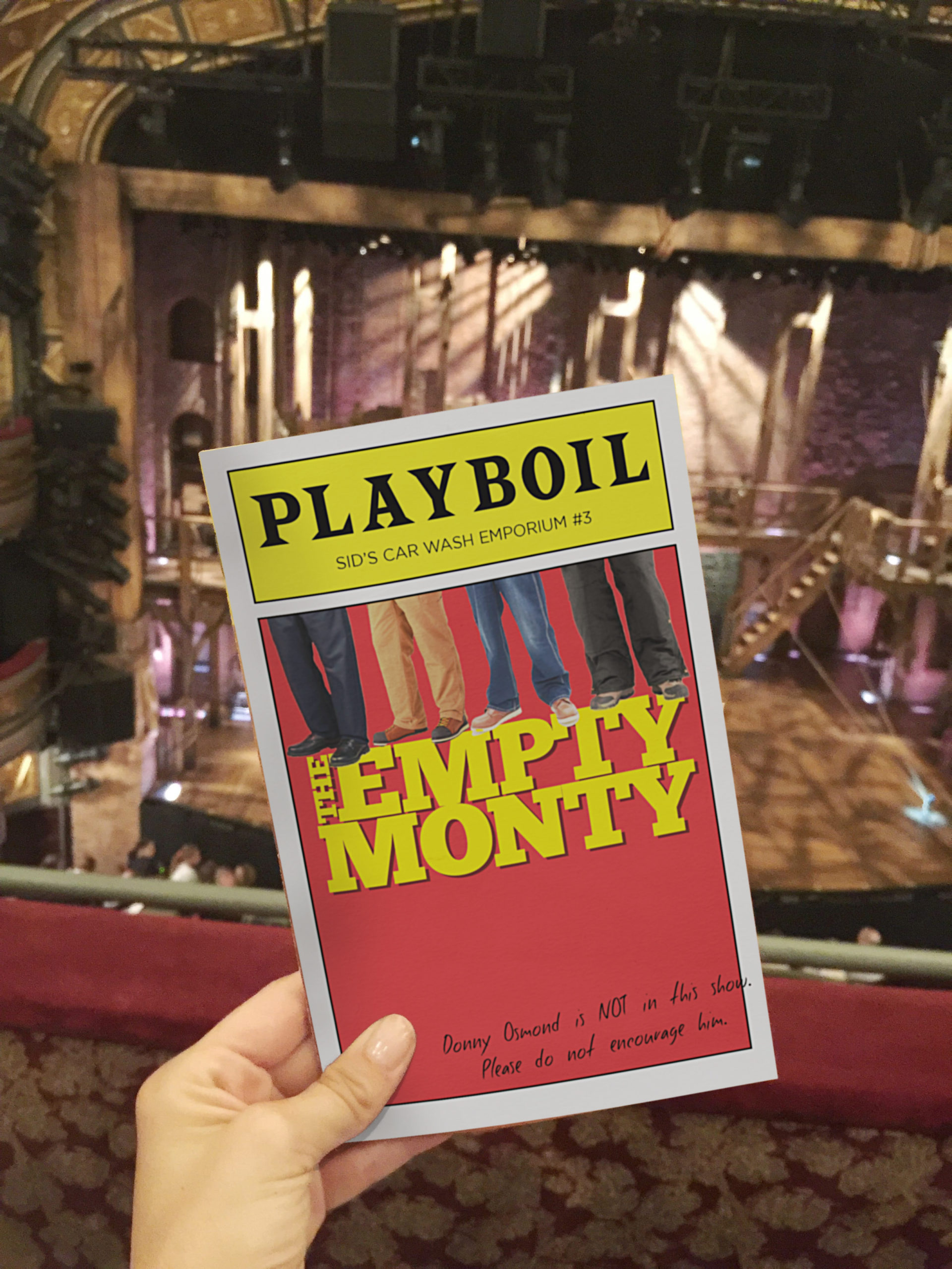 A hand holding the program for "The Empty Monty." A stage can be seen in the background.