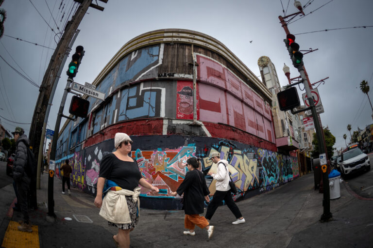 An abandoned building on a corner in the Mission District of San Francisco. It is covered in graffiti.Pedestrians walk by in the foreground.