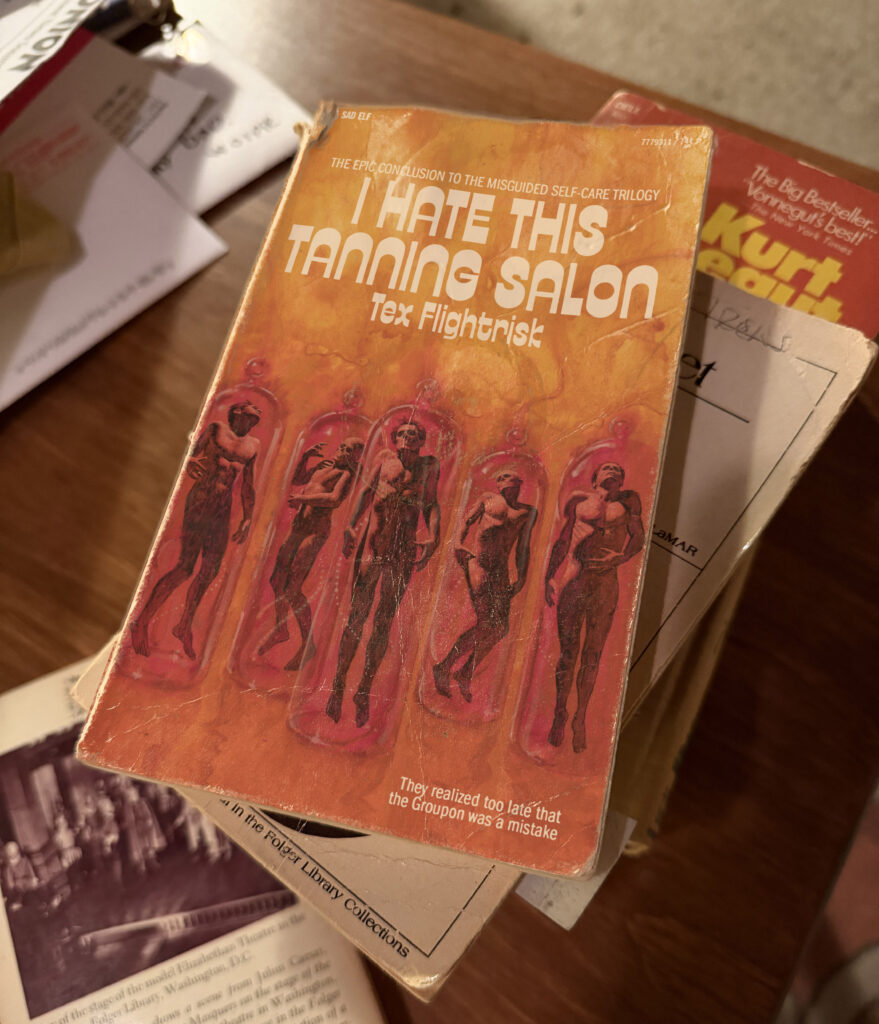A pile of books on a table. The top book's cover has a painting of decaying human bodies in tubes. The book is "I Hate This Tanning Salon" by Tex Flightrisk.